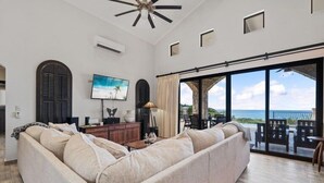 A living room with a large beige sectional sofa, a flat-screen TV, a ceiling fan, and a view of the ocean through large sliding glass doors.
