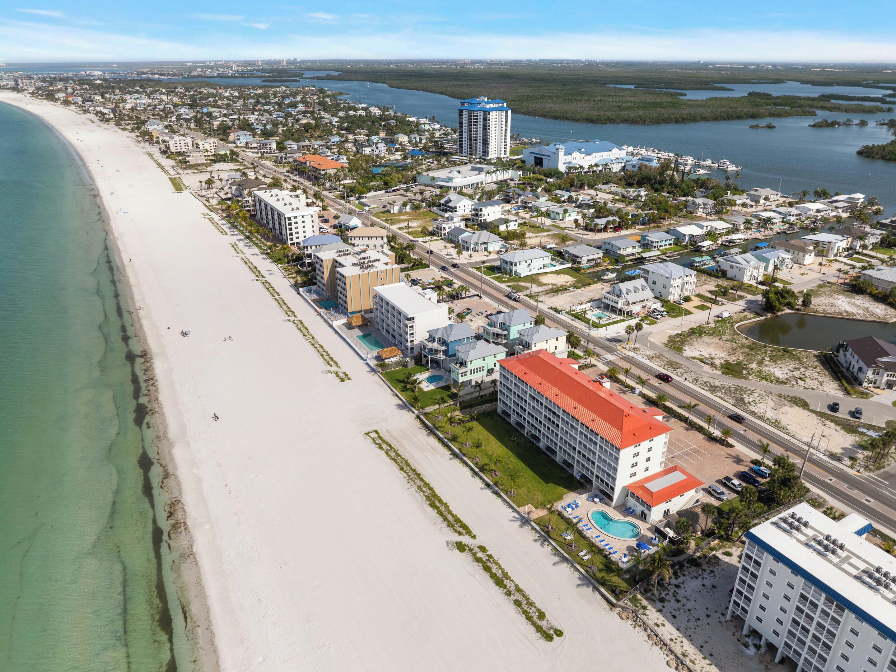 Ett flygbildsbild av en kuststad med en strand, en småbåtshamn och olika byggnader, inklusive hotell.