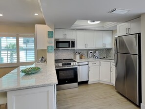A modern kitchen with white cabinets, stainless steel appliances, a large island, and a view through the window. - Pointe South 107 | Stunning Views + Resort Pool (Fort Myers Beach)