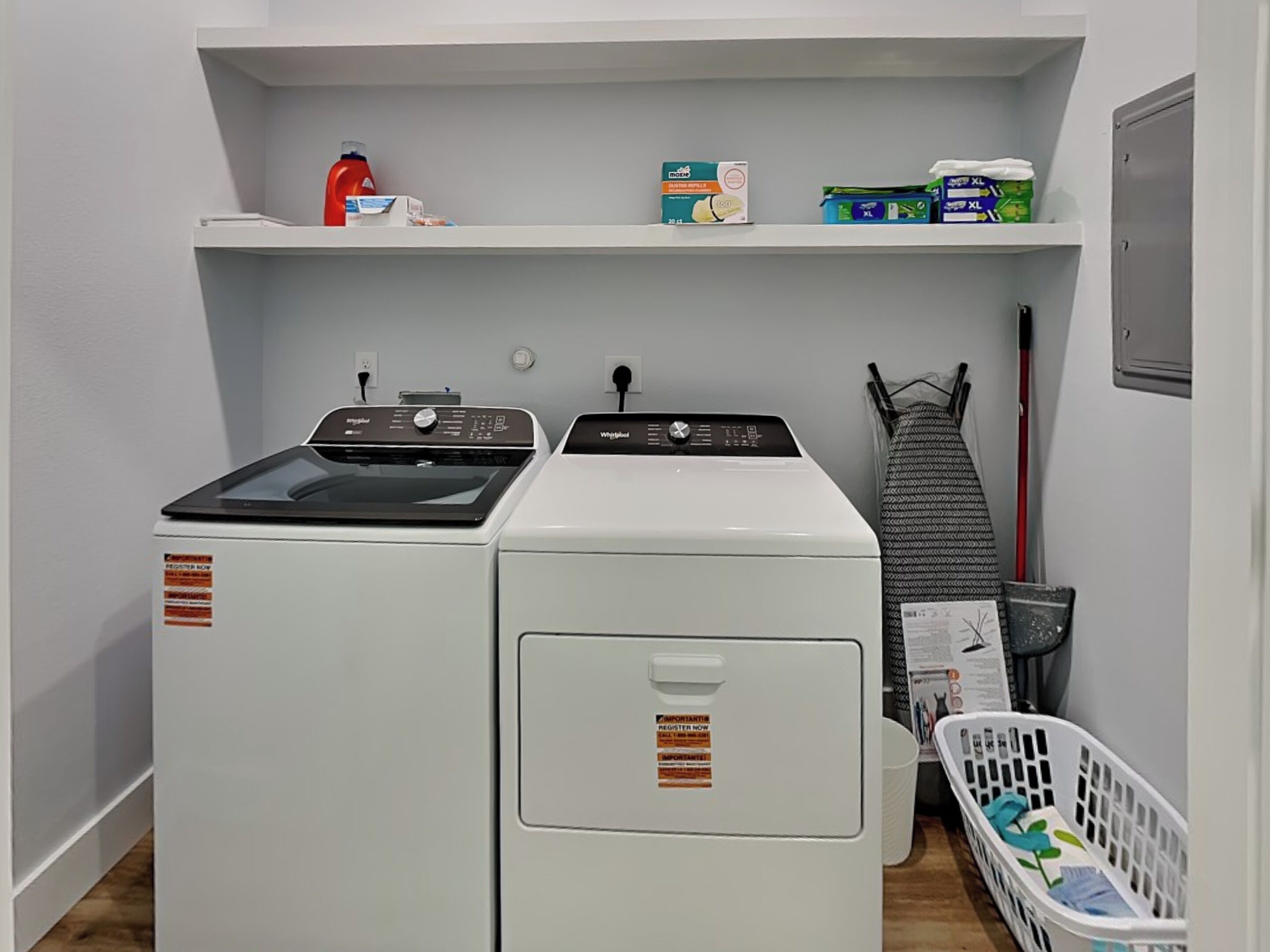 Une buanderie équipée de deux machines à laver et deux sèche-linge à chargement frontal, d’une étagère contenant des produits de nettoyage, d’un panier à linge et d’une serpillière.