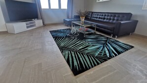 Ein Wohnzimmer mit Flachbildfernseher, einem dunklen Sofa, einem Glastisch, einem gemusterten Teppich und einem Fenster mit Vorhängen.
