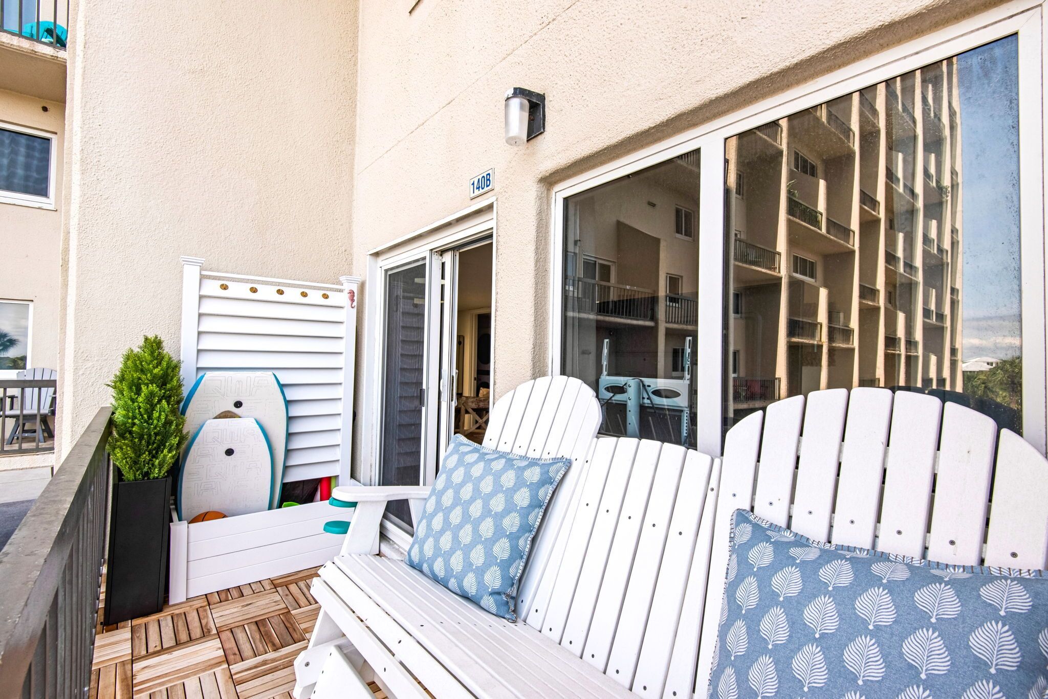 A balcony with a white Adirondack chair, a small potted plant, and a window with a view of a building.