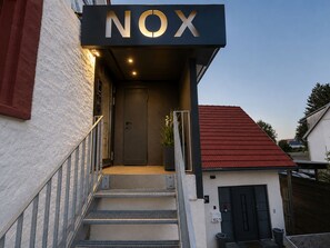 A modern building entrance with a 'NOX' sign, a staircase, and a small adjacent structure with a red roof. - NOX 3 Tokyo, Deluxe Double Room, 2 Beds, Bathroom, Minibar, Digital Check-In - No Reception (Walzbachtal)