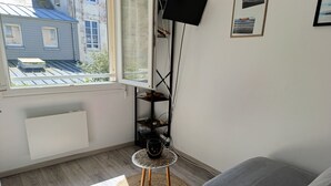 Ein Zimmer mit grauem Sofa, einem kleinen runden Tisch mit Geflecht, einem Fernseher auf einem schwarzen Schrank und einem Fenster mit Blick auf Gebäude.