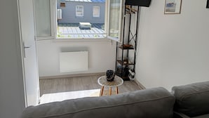 Ein Zimmer mit einer Couch, einem Fernseher auf einem Schrank, einem Fenster mit Blick auf ein Haus und einem kleinen Tisch mit einem dekorativen Gegenstand.