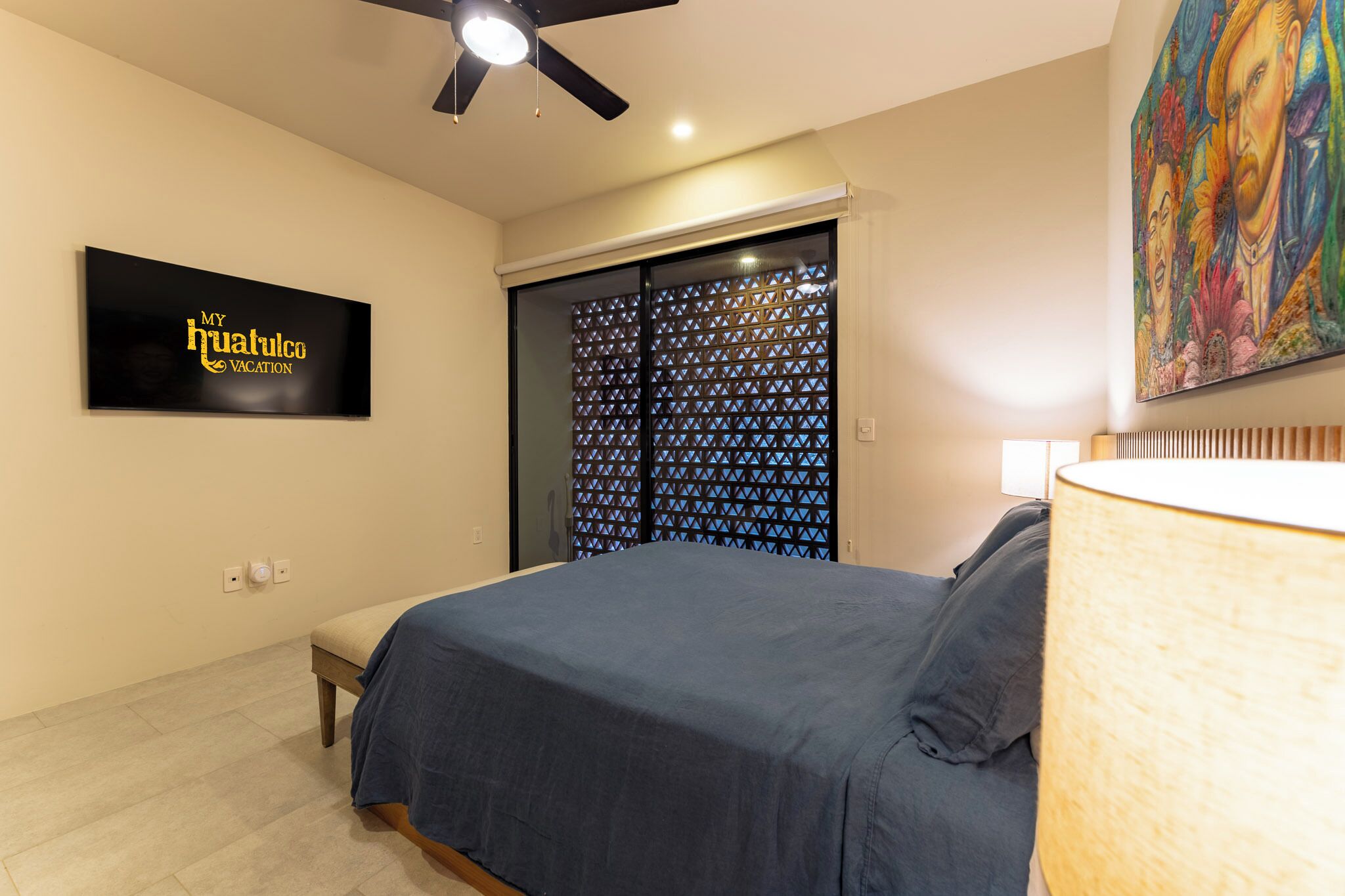Una camera da letto con un letto, un ventilatore a soffitto, una TV, un quadro e una finestra con persiana decorata.