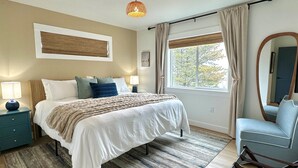 A bedroom with a bed, bedside tables, a chair, a window with curtains, and a hanging light fixture. - Villa w/ Spa, Theater, Arcade & Hot Tub · Pets OK (Ocean Shores)