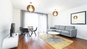 Ein modernes Wohnzimmer mit einem grauen Sofa, einem weißen Couchtisch und einem gelben Teppich. Zwei schwarze Sessel, ein Fernseher und ein gerahmtes Bild sind ebenfalls vorhanden.
