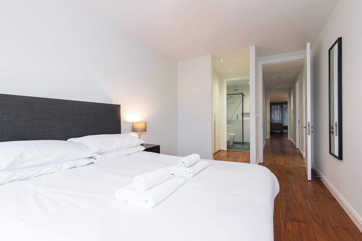 Quarto de hotel com cama bem arrumada, mesa de cabeceira com abajur e corredor que leva a um banheiro.