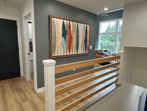 A modern interior with a wooden staircase, a gray wall with a colorful abstract painting, and a window with a view of greenery.