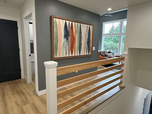 Un intérieur moderne avec un escalier en bois, un mur gris orné d’un tableau abstrait aux couleurs vives, et une fenêtre donnant sur un paysage verdoyant.