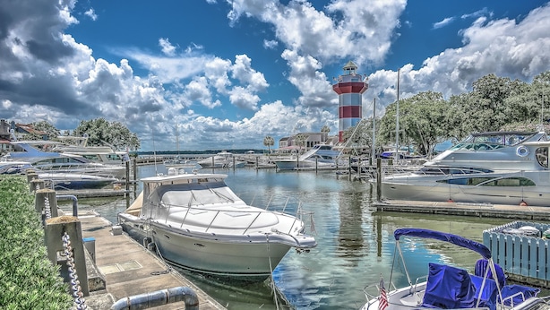 Un puerto deportivo con barcos amarrados, un faro al fondo y cielo despejado.