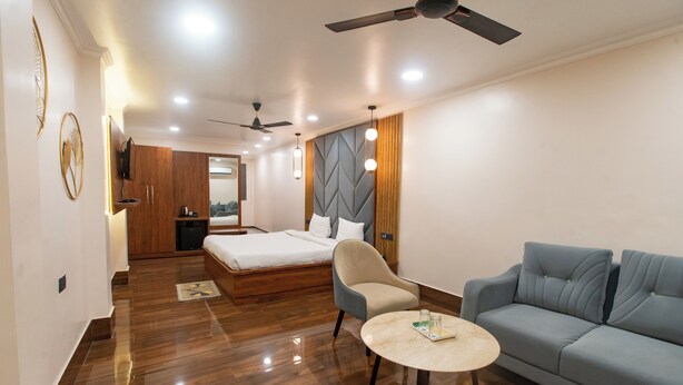 Habitación de hotel con cama, sofá, sillón, una pequeña mesa, ventilador de techo y televisor montado en la pared.