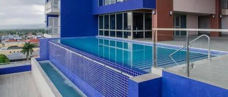 A modern building with a blue exterior, a swimming pool, and a view of the cityscape.