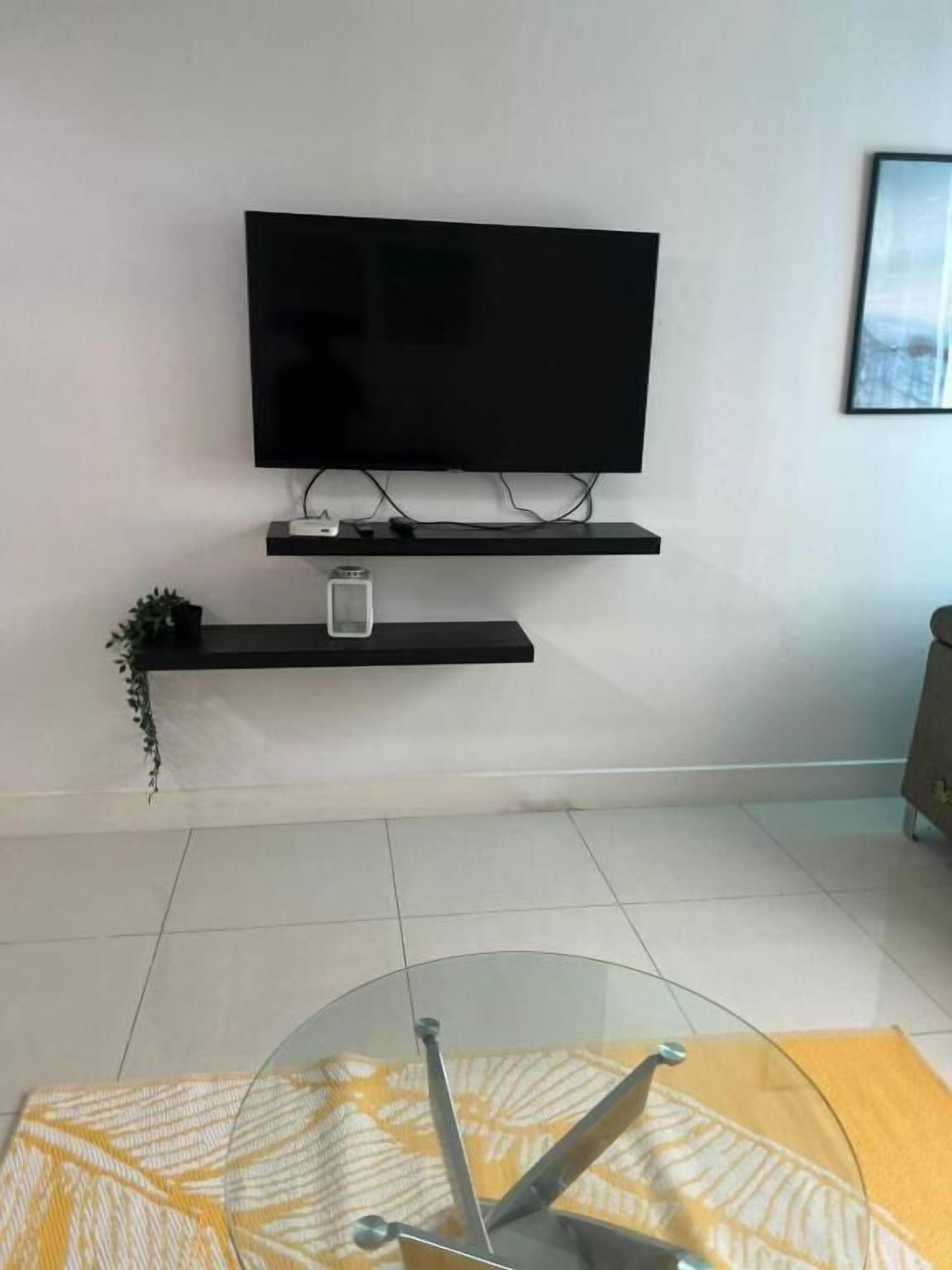 A modern living room with a flat-screen TV mounted on the wall, a small plant, and a glass table with a yellow patterned tablecloth.