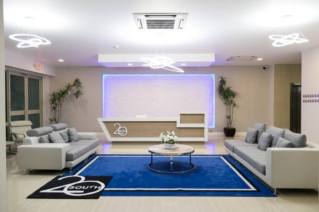 A modern lobby with a reception desk, sofas, a coffee table, and a blue carpet with a logo.