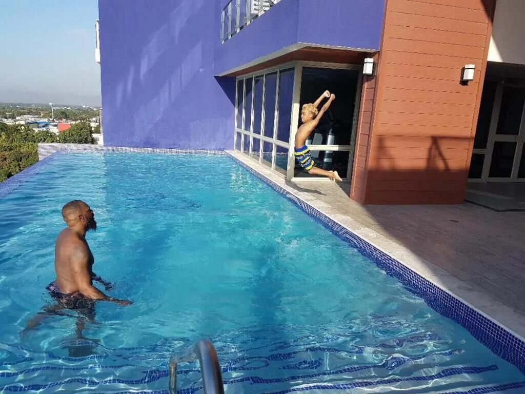 Une piscine avec une personne qui plonge, un bâtiment à la façade violette et un ciel dégagé.
