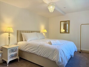 A neatly made bed with white linens, two bedside lamps, a nightstand with decorative drawers, a gold-framed mirror, and a chandelier.