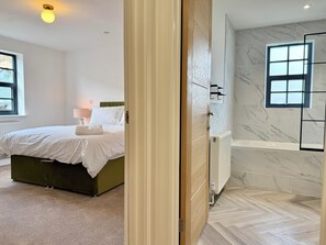 Ett hotellrum med en säng, ett sängbord med en lampa, ett element och ett badrum med badkar och väggar klädda i marmor.