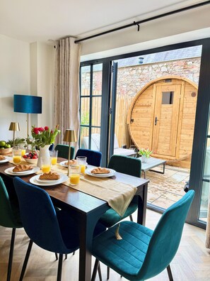 A dining area with a table set for breakfast, including croissants and orange juice, and a view of a wooden building outside. - Luxury/Hot tub/sauna/Sleeps 9/ Parking (Chepstow)