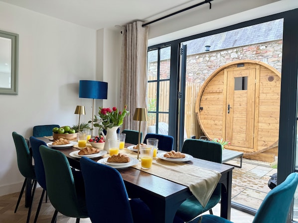 A breakfast table set with croissants, fruit, and juice glasses, a vase with flowers, and a large wooden door in the background.