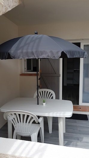Eine Terrasse mit einem weißen Tisch und Stühlen, einem dunklen Sonnenschirm und einer Glasschiebetür zu einem überdachten Bereich.