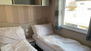 Huoneessa on kaksi erillistä sänkyä, ikkuna, jossa on verhot, seinälle kiinnitetty hylly ja pistorasia.