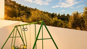 A swing set with two swings, a concrete ground, and a white wall with a view of a forested hillside. - OASIS 360° Apartment (Kos)