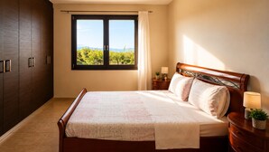 A bedroom with a bed, a nightstand, a lamp, a window with a view of greenery, and a wardrobe. - OASIS 360° Apartment (Kos)