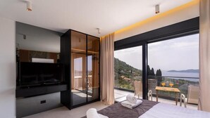 Quarto de hotel moderno com uma cama grande, televisão de tela plana, varanda com vista para o mar e área de jantar com mesa e cadeiras.