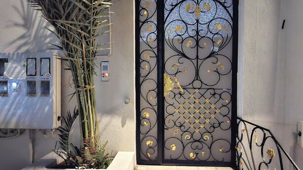 Una entrada con reja metálica decorativa, una jardinera de concreto con una planta alta y un conjunto de escalones de piedra que conducen a la entrada.