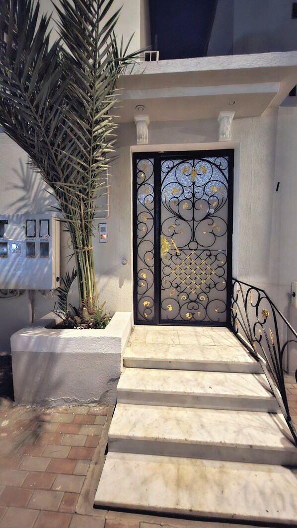 A gated entrance with a decorative metal gate, a concrete planter with a tall plant, and a set of stone steps leading up to the gate.