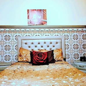 Un dormitorio con cabecera capitonada, papel tapiz estampado y cama con ropa de cama floral y almohada con estampado de rosas.