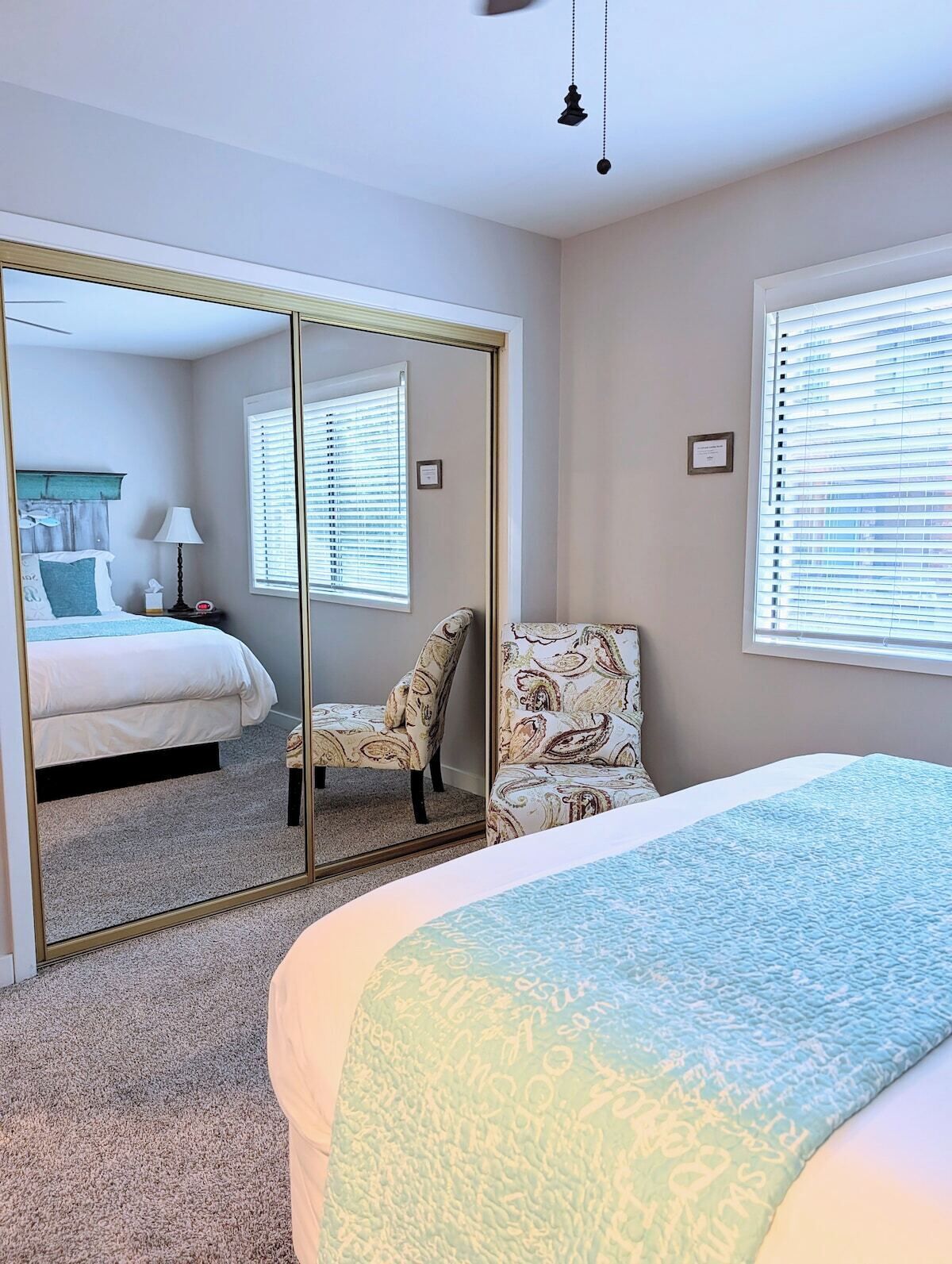 A bedroom with a bed, a chair, a mirror, and a window with blinds.