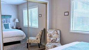A bedroom with a bed, a chair, a mirror, and a window with blinds.