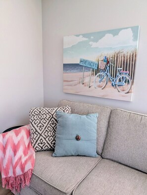 A room with a beige sofa, a blue cushion, and a wall-mounted painting of a beach scene with a bicycle. - Lake Breeze Retreat · Newly Remodeled · Lake Views (Chelan - Beach Bungalows)