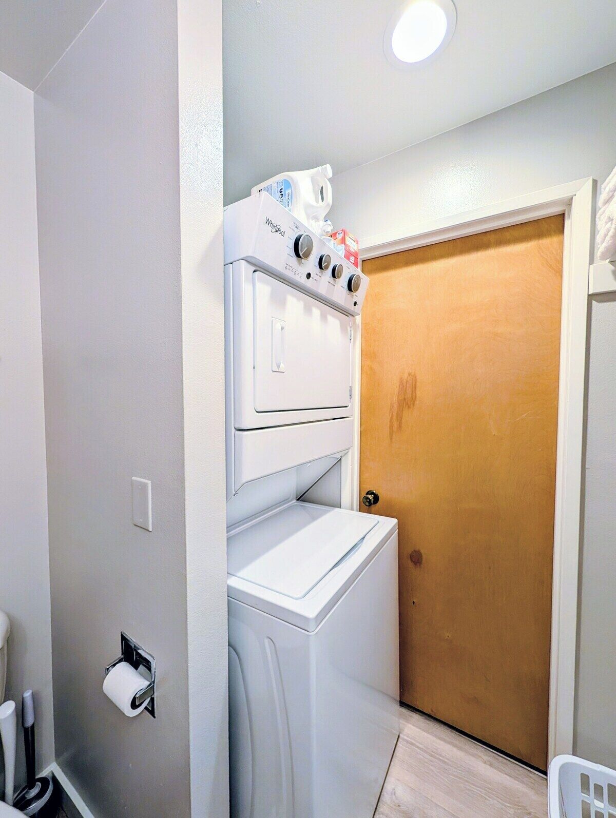 A laundry room with a stacked washer and dryer, a wooden door, and a white wall.