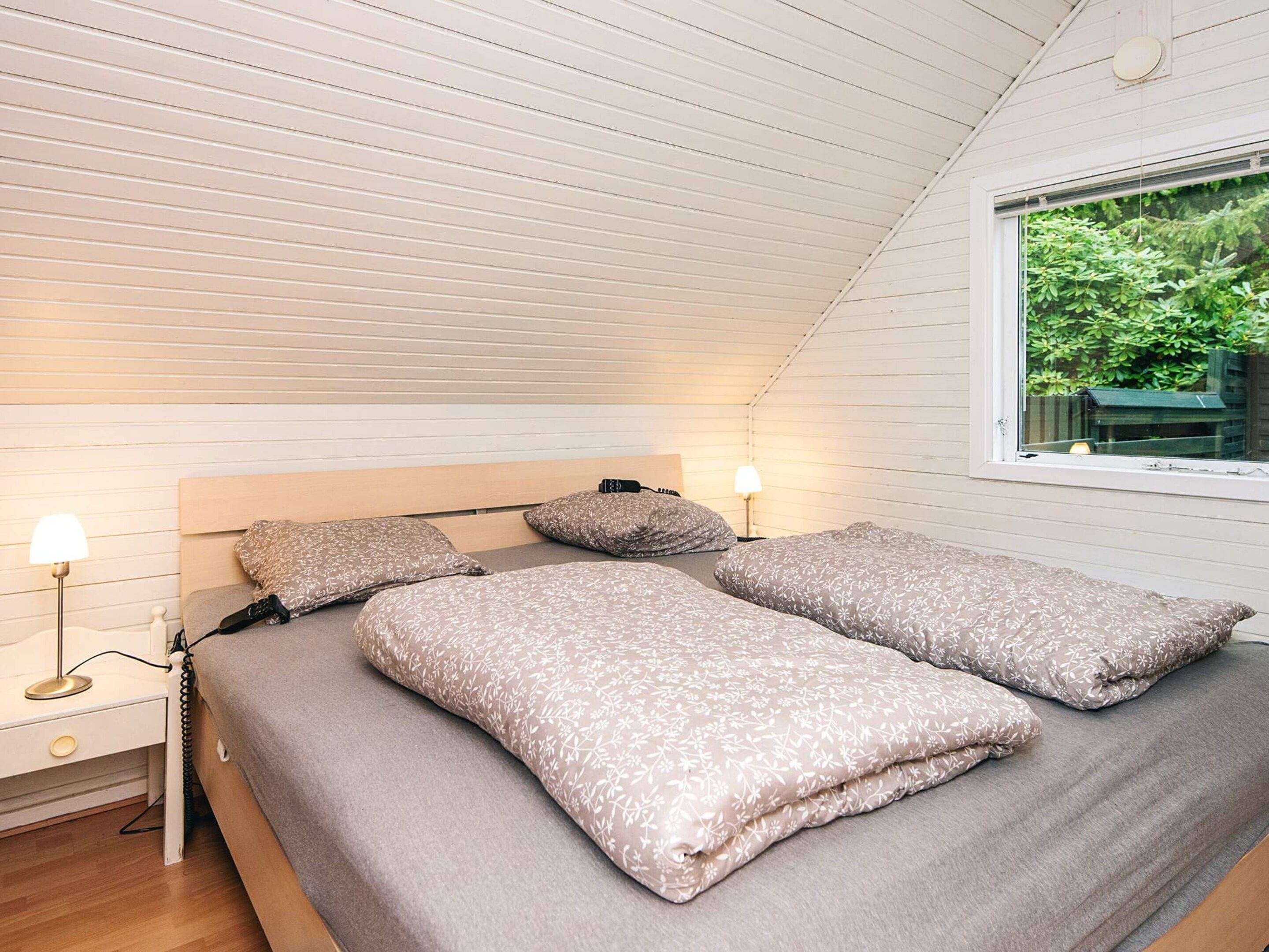 Une chambre avec un plafond en pente, un lit recouvert d’une literie à motifs floraux, des lampes de chevet et une fenêtre donnant sur un paysage verdoyant.