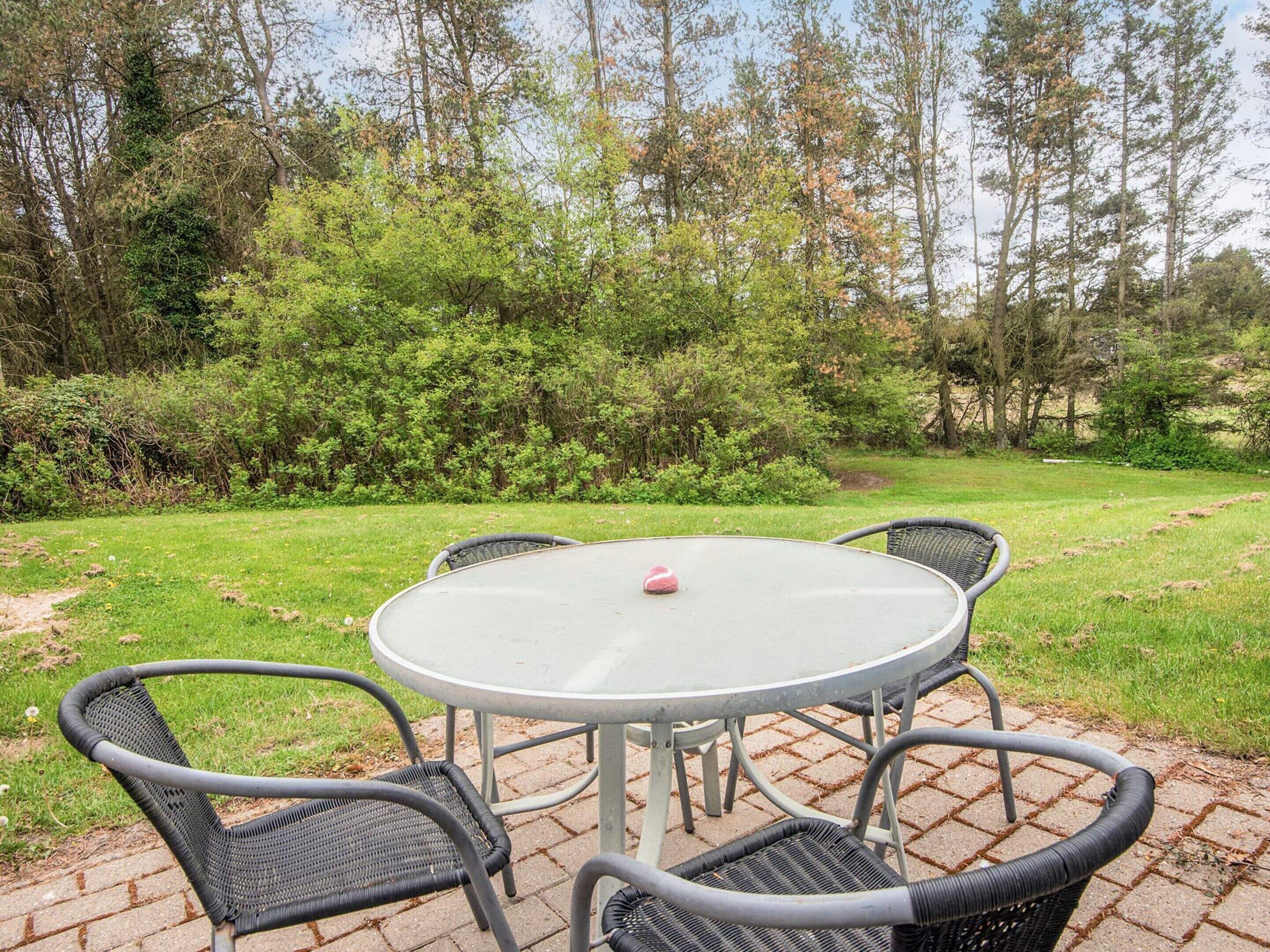 Ein runder Gartentisch mit vier Stühlen, umgeben von einer Pflasterterrasse, mit einer Rasenfläche und Bäumen im Hintergrund.