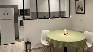Une cuisine moderne avec des meubles blancs, un four à micro-ondes et un réfrigérateur. On y trouve une table à manger recouverte d’une nappe verte et deux chaises. Un miroir est accroché au mur, au-dessus de la table.