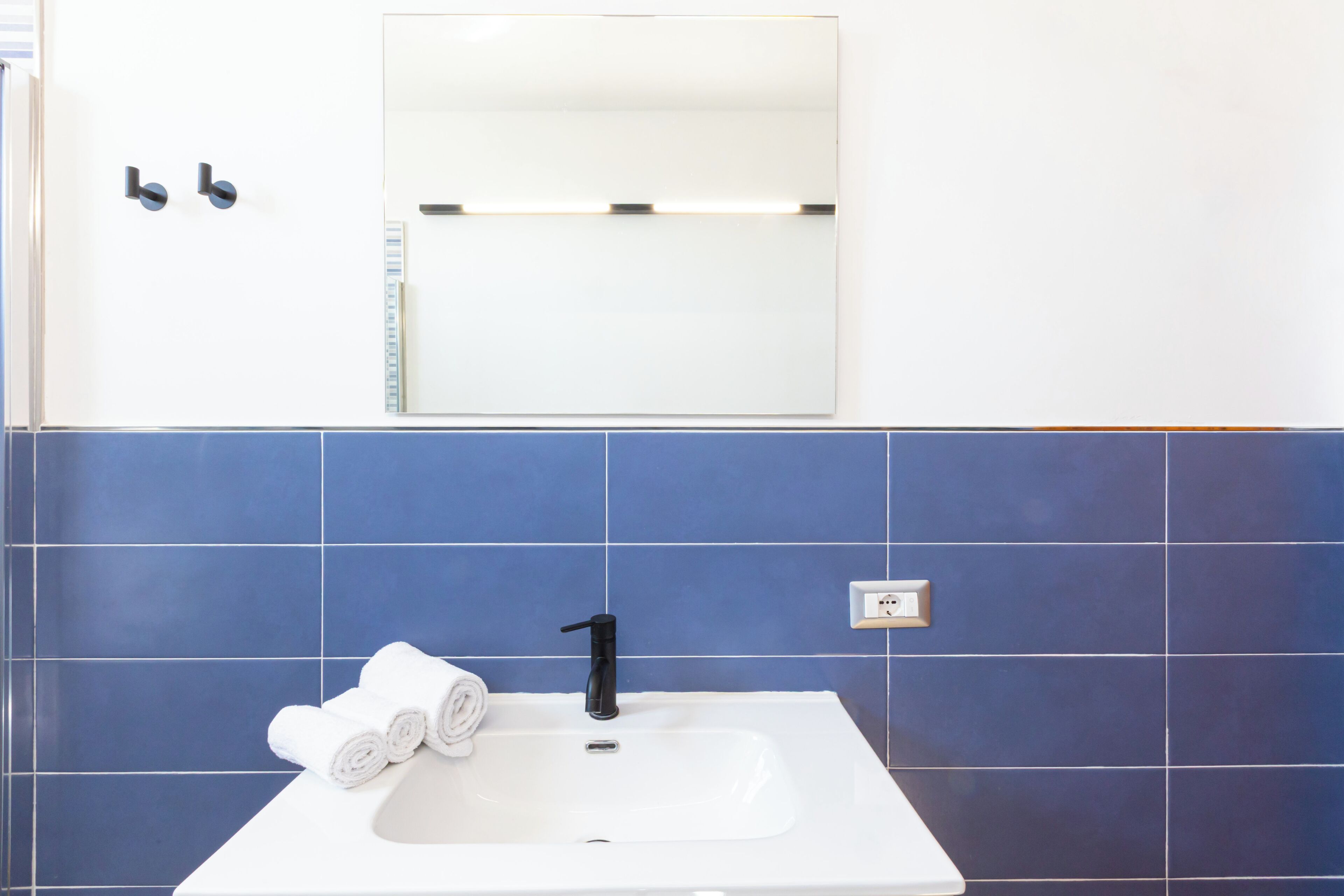 A bathroom with a white sink, blue tiles, a mirror, and two black towel holders.