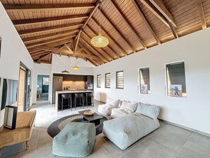 A spacious living area with a high wooden ceiling, a kitchen with a central island, and a dining area with bar stools. - LA PETITE VILLA 2CH PISCINE PROCHE MER (Sainte-Anne)