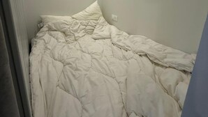 Een kleine, zwak verlichte kamer met een enkel bed, een stopcontact en een ventilatie rooster.