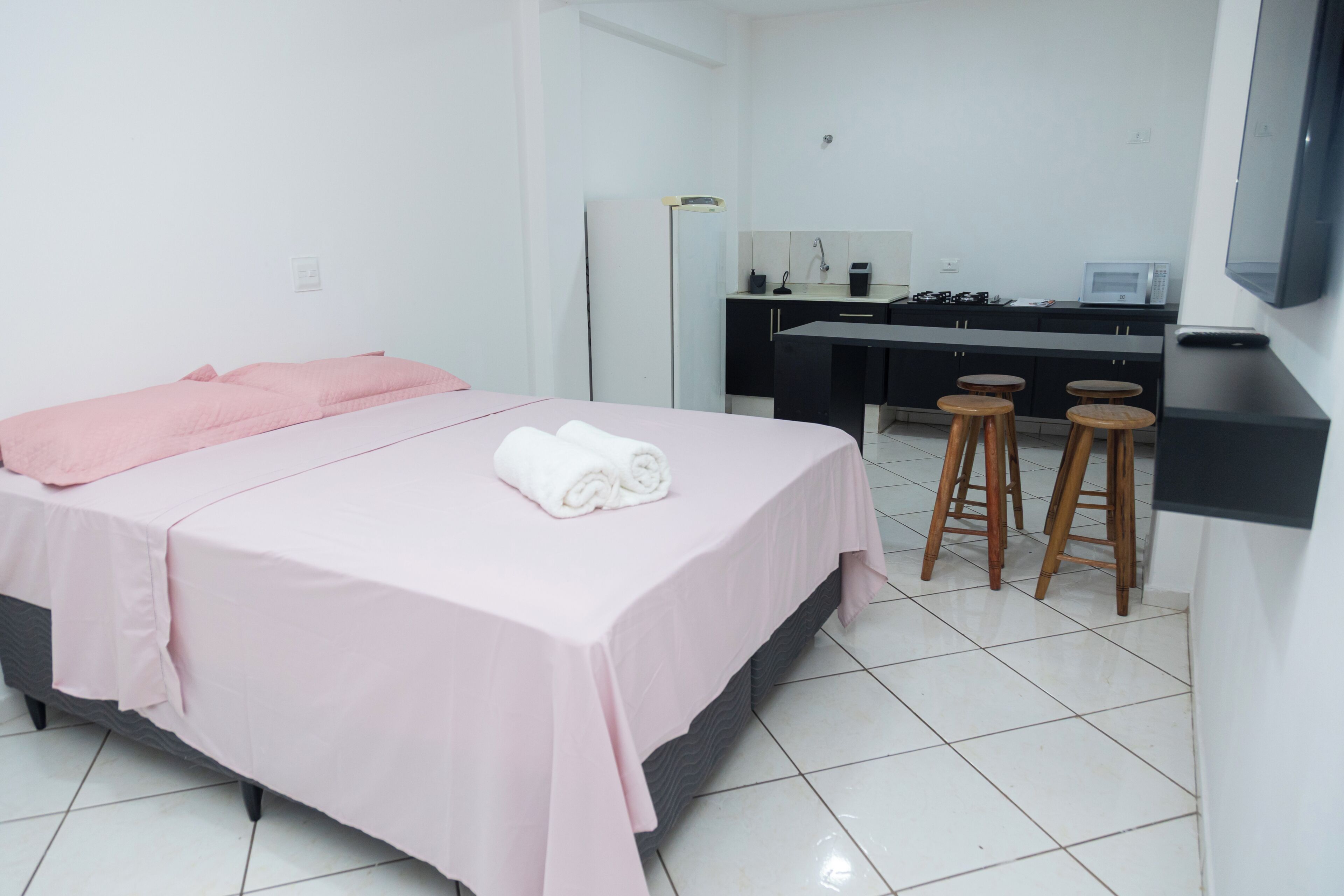 A hotel room with a bed, a desk, a kitchen area, and a TV mounted on the wall.