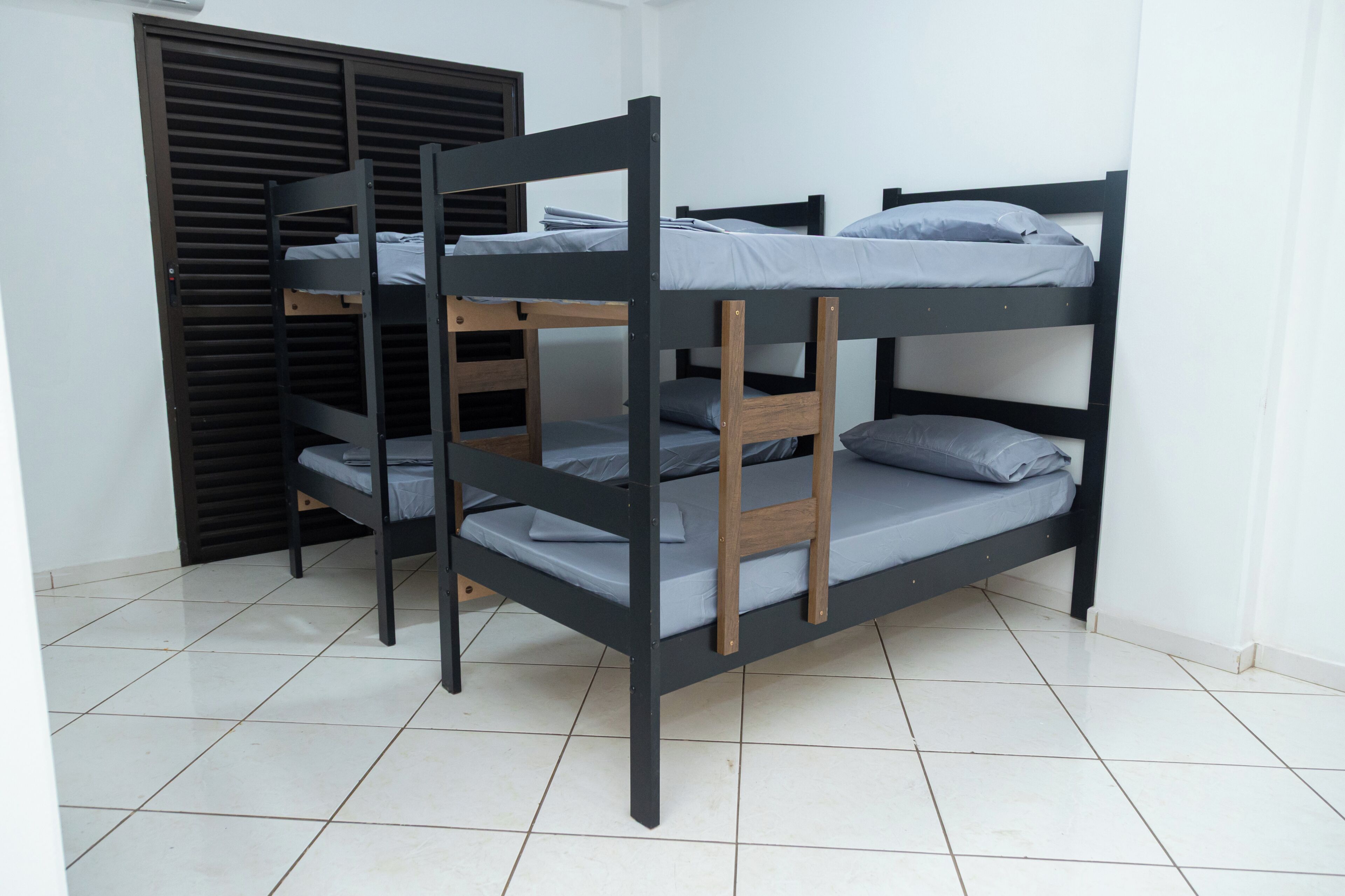 Uma cama beliche com estrutura de madeira e roupa de cama cinza, num quarto com pavimento em azulejo branco e um armário de cor escura.