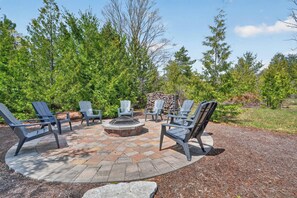 A circular outdoor seating area with Adirondack chairs and a fire pit, surrounded by a gravel path and dense greenery.