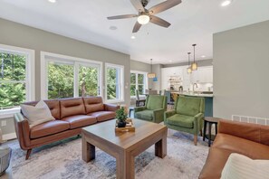 A living room with a brown leather sofa, a wooden coffee table, and green armchairs. There is a ceiling fan, large windows, and a view of the kitchen.