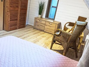 Quarto com piso de madeira, cama com colcha estampada, cômoda de madeira, cadeira de vime e janela com vista.
