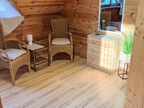 Interior acolhedor de uma cabana de madeira, com duas cadeiras de vime, uma mesa pequena, um aparador e um espelho.