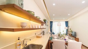 A modern kitchen with a wooden shelf, a sink, and a dining area with a table and chairs. - A la Carte - Coralie Luxury Seaview Aprtment (Đà Nẵng)